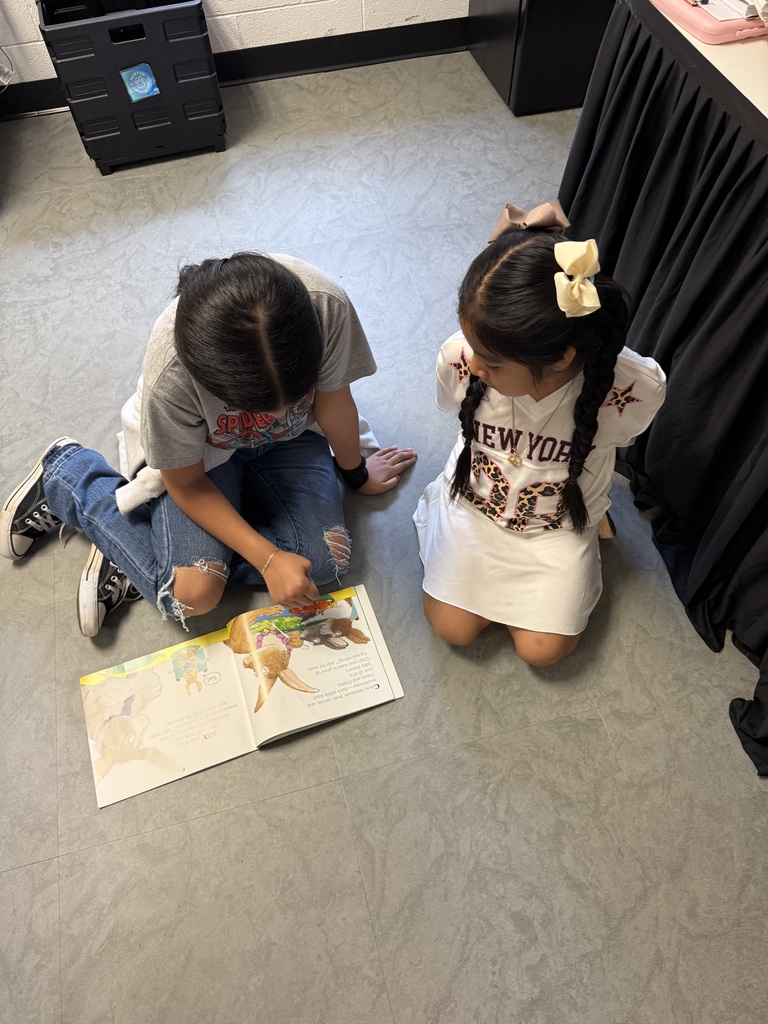 Two children sit on the floor, one reading a book aloud to the other. One wears a white dress, and the other, a gray shirt.
