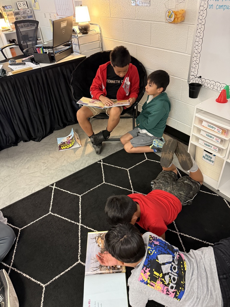 Four children read books on a patterned rug in a classroom with white walls and furniture.
