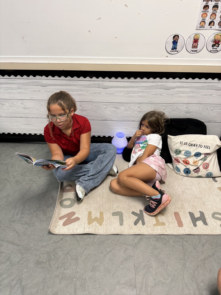 Two children sit on a floor, reading. One holds a book, the other wears glasses. A pillow, lamp, and rug with letters are nearby.