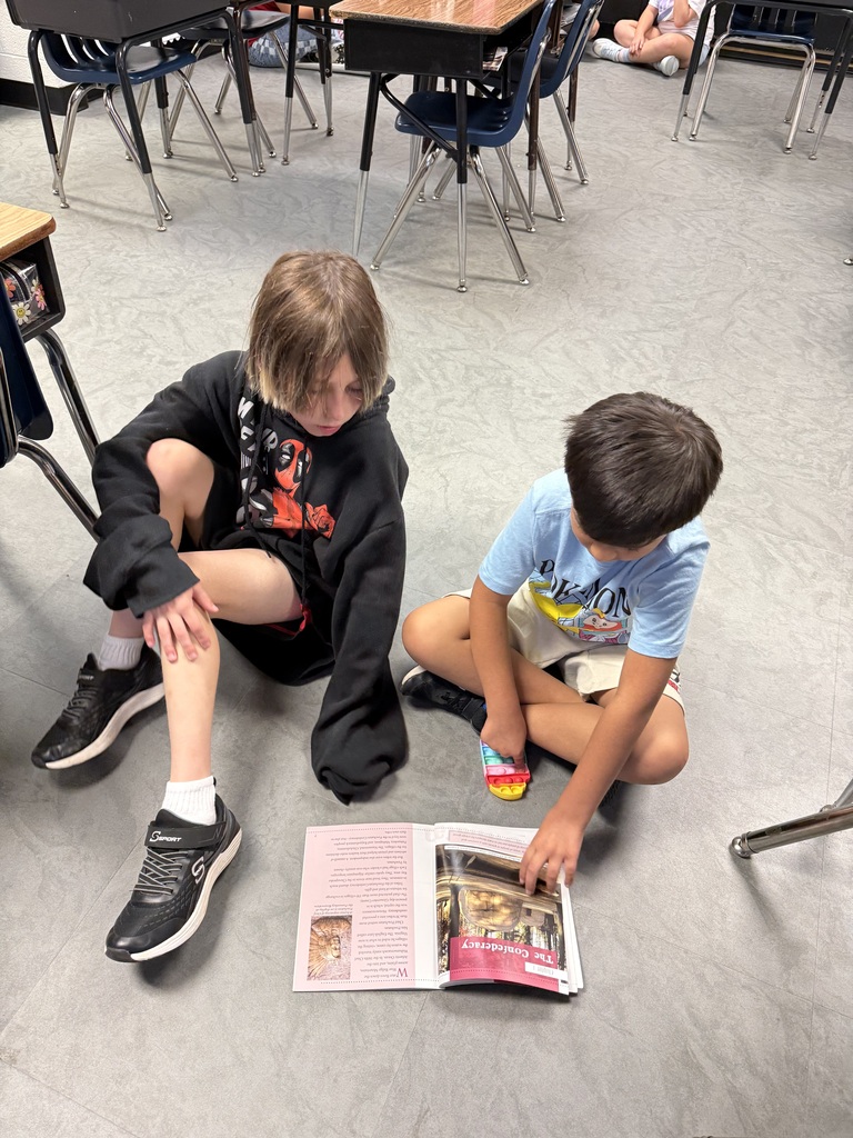 Two children sit on a classroom floor, one reading a book. A table with chairs is in the background.