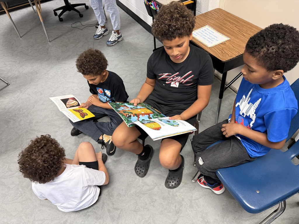 Four children sit on the floor, reading books. Two read aloud, while two listen. They sit in a classroom.
