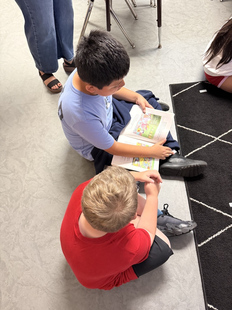 Two children sitting on the floor reading a book, one wearing a blue shirt and the other a red shirt.