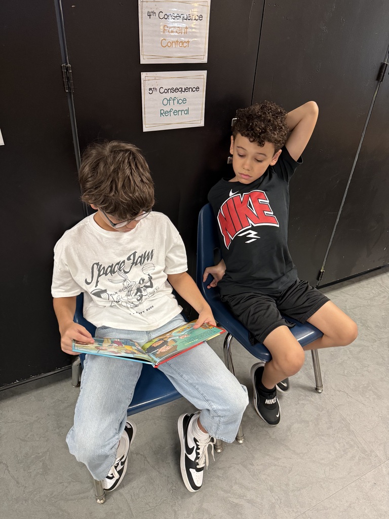 Two boys sit on blue chairs, one reading a book and the other leaning back. Behind them is a black wall with signs.