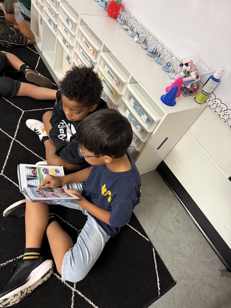 Two children sit on the floor reading a book. They wear glasses and sneakers. A white shelf with drawers and items behind them.