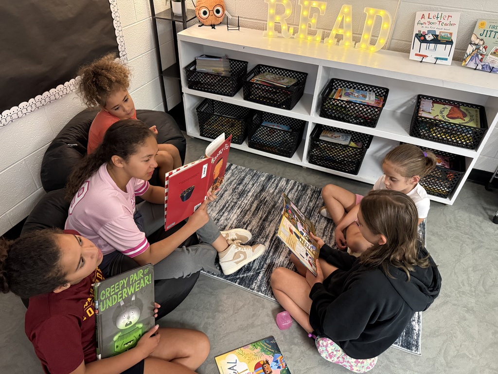 Five children sit on the floor in a classroom. One reads a book to others, with shelves and storage boxes in the background.