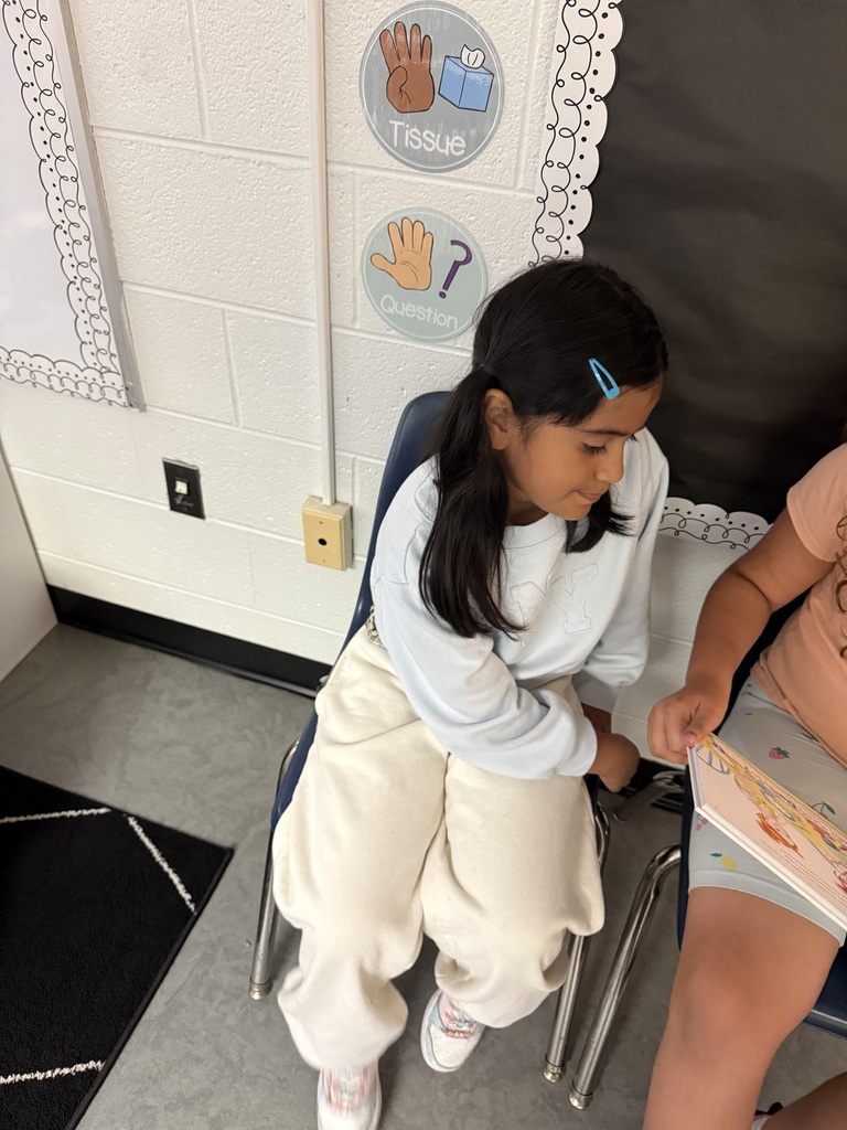 Two students in a classroom, one seated and reading a book, the other standing, both wearing casual clothing.