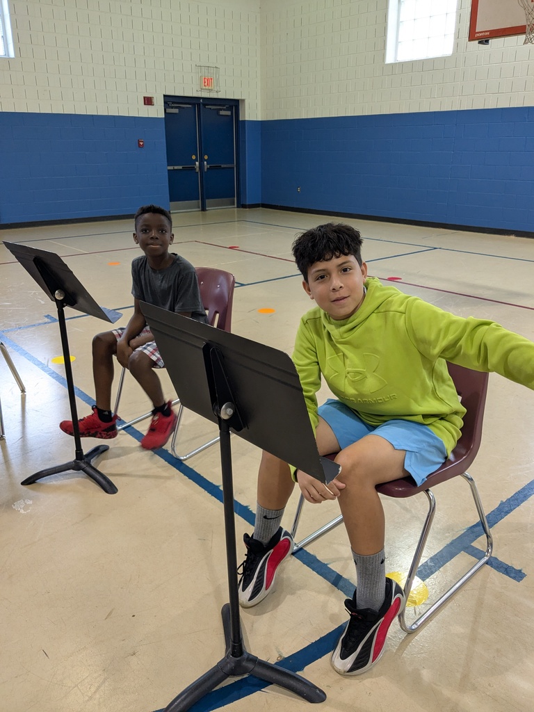 Two boys seated in chairs, one reading music. A music stand is between them. Behind them, a gym with blue walls and a door.