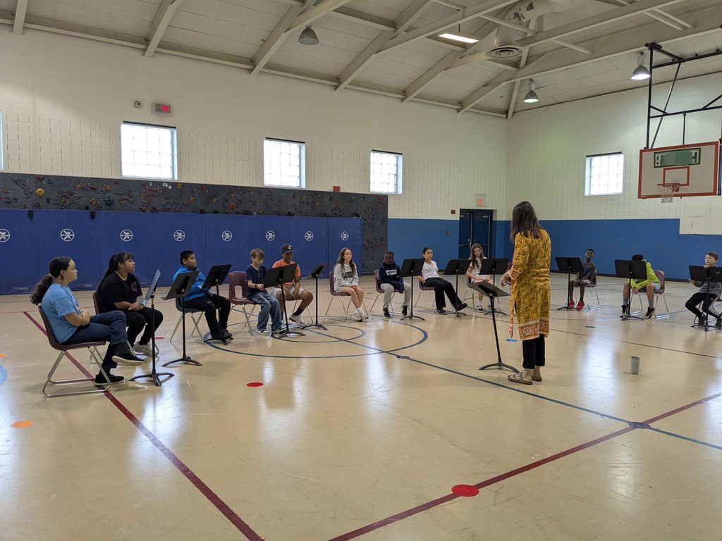 A group of individuals seated in chairs, listening to a woman with a microphone in a basketball court.