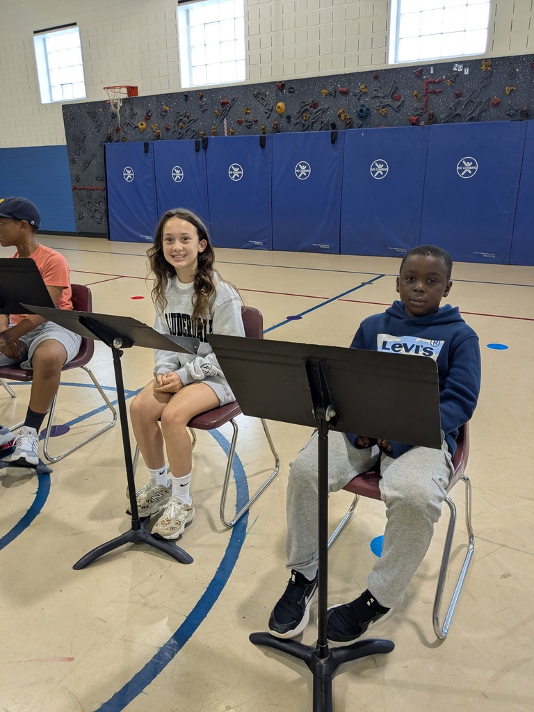 Three students sit in chairs in a gym with musical instruments. The background includes a climbing wall and windows.