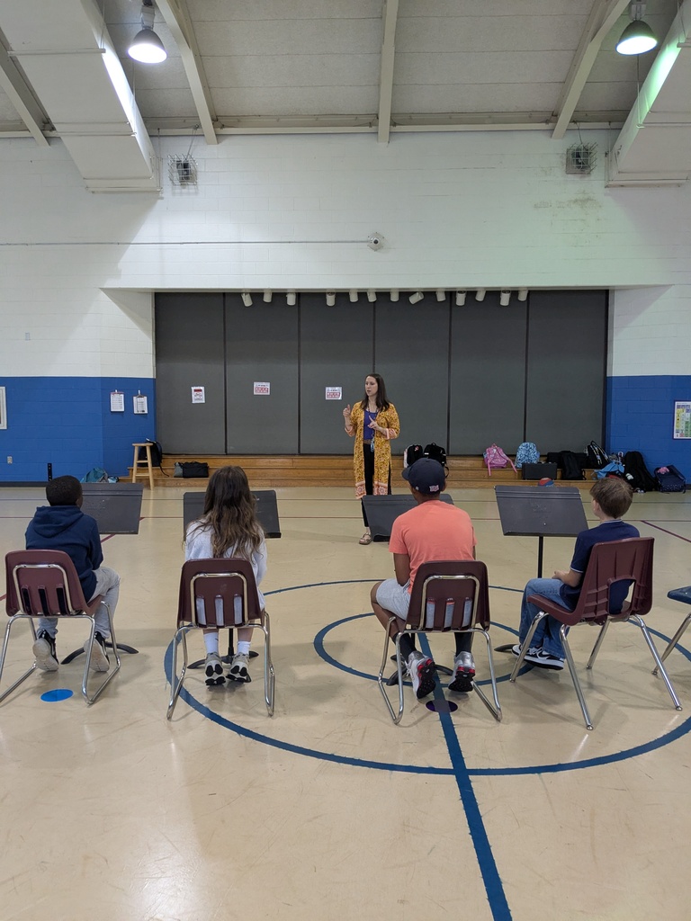 A woman stands in front of four students, who sit in a circle on chairs. The group is in a room with a blue line on the floor and a gray wall.