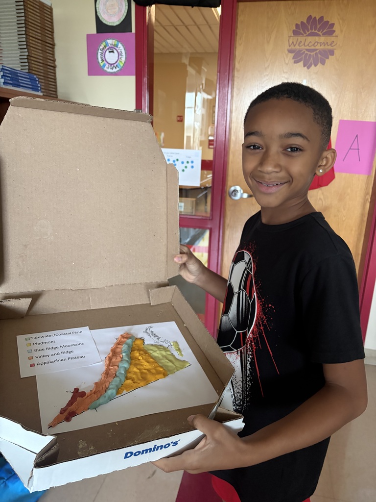 A smiling student holds an open pizza box displaying his completed 3D clay map of Virginia’s geographic regions.