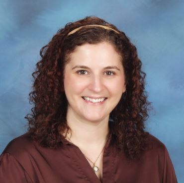 A smiling woman with curly hair wears a maroon shirt. She has a headband and a necklace.