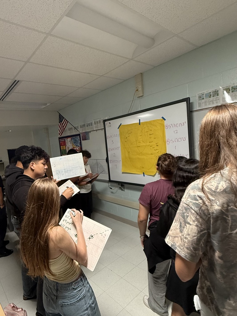 Several students stand in a classroom around a whiteboard with yellow paper and notes, with one student writing on paper.