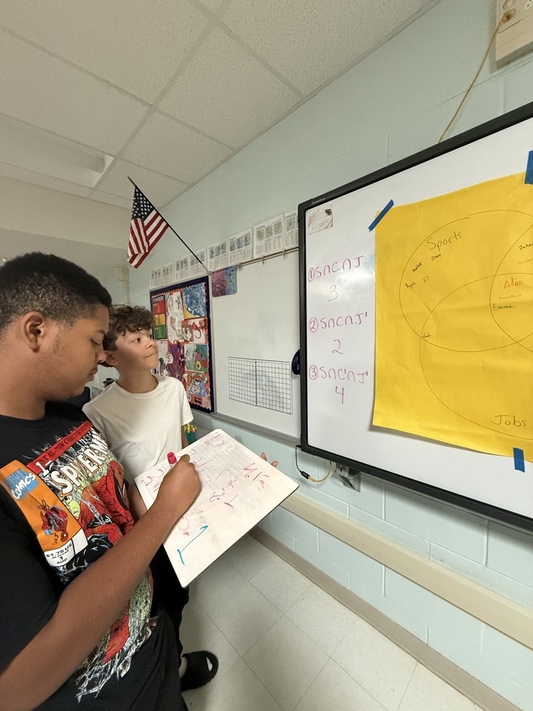 Two boys in a classroom looking at a large paper. One is writing on a smaller piece of paper.