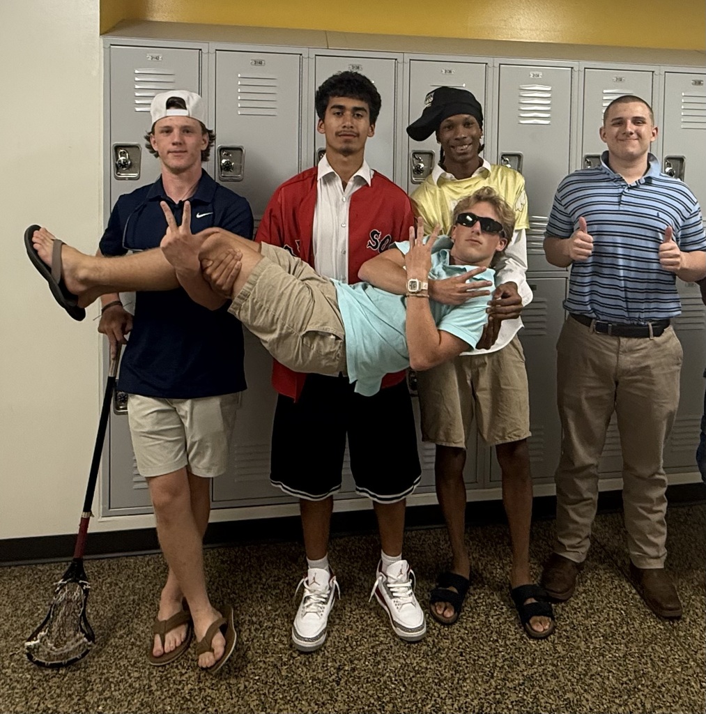 Five people in a locker room, one person lying down with a lacrosse stick, others standing.