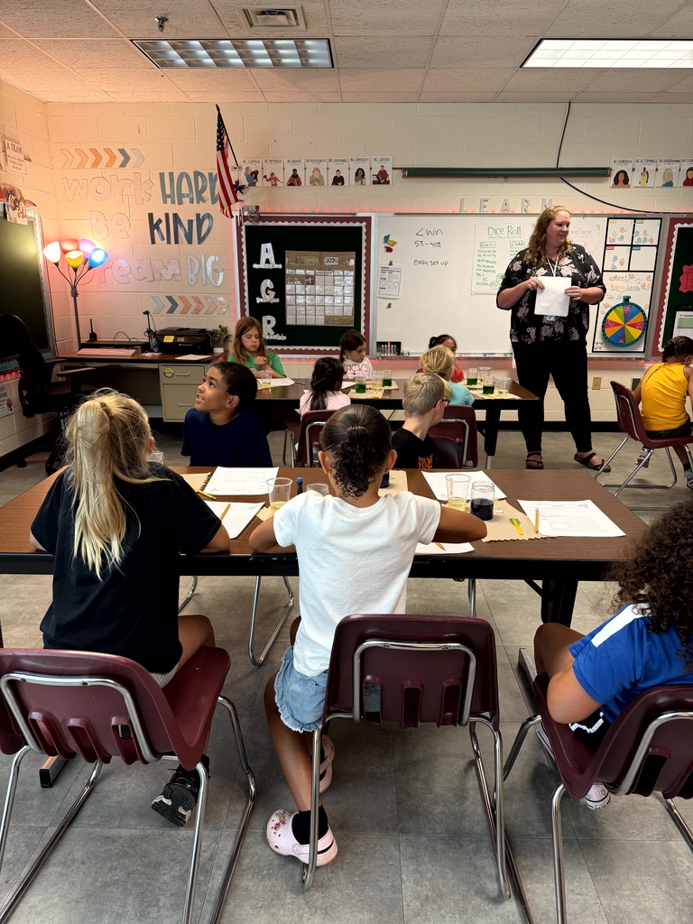 A classroom with children seated at tables. A teacher stands at a table, facing the children, and holds a paper. Whiteboards and classroom decor are in the background.