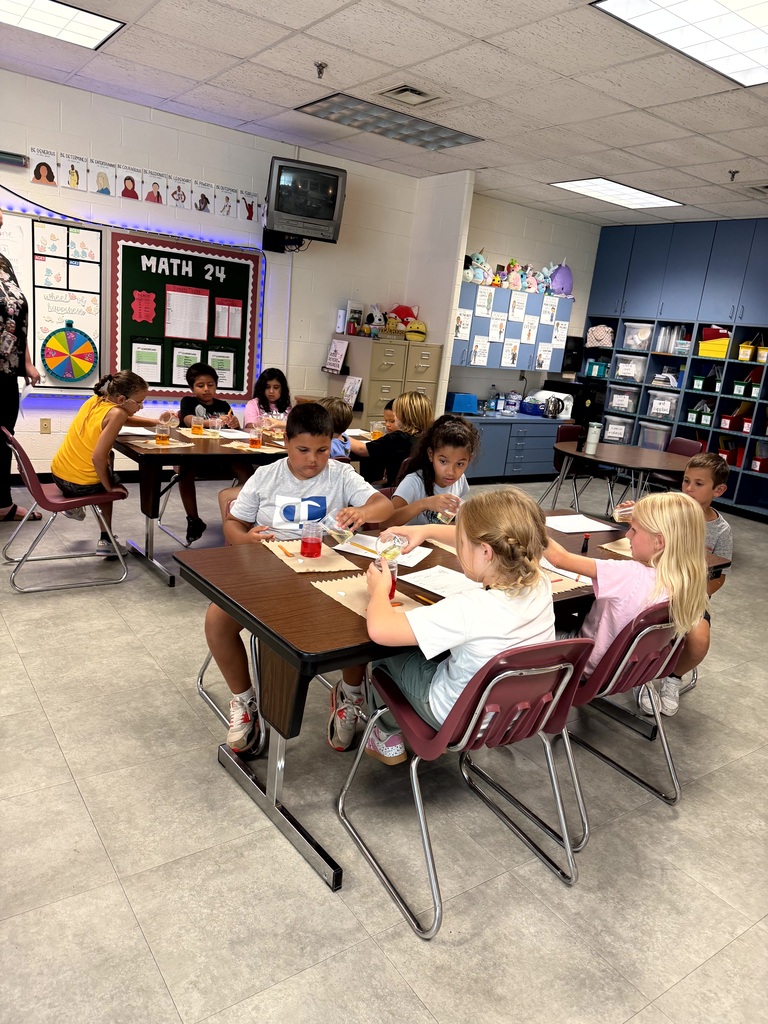 Students seated around desks in a classroom, one wearing a white shirt, another in a yellow shirt.