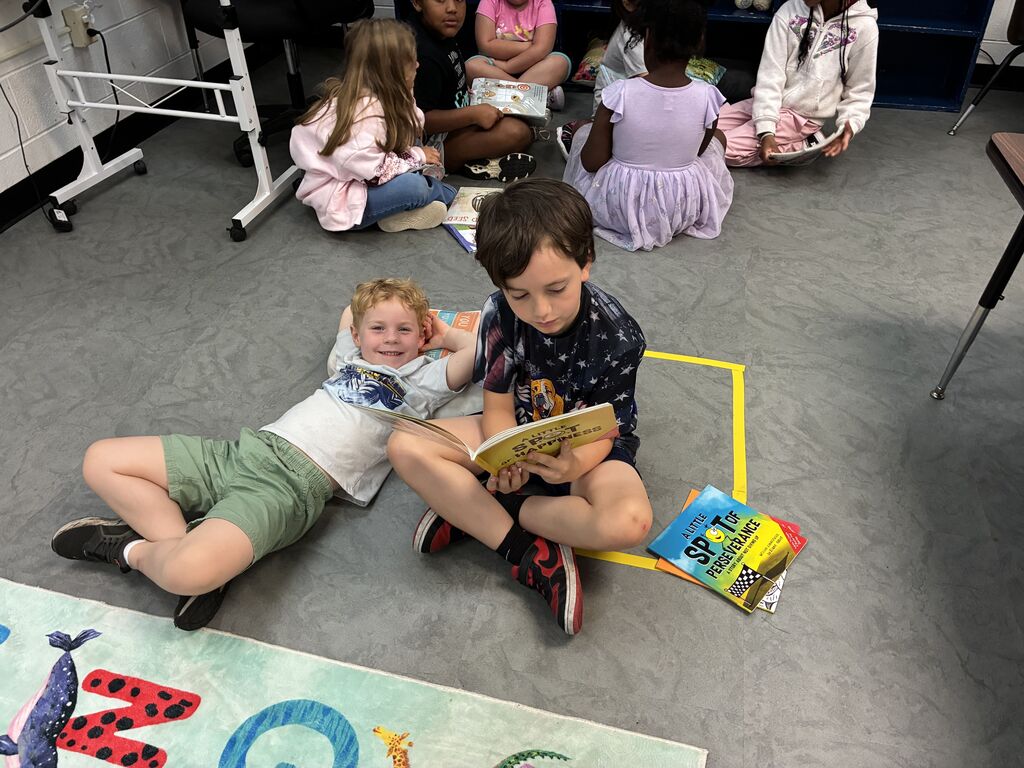 Kids sit on floor, a few with books, one reading aloud. Colorful rug on floor. Chairs and a table in background.
