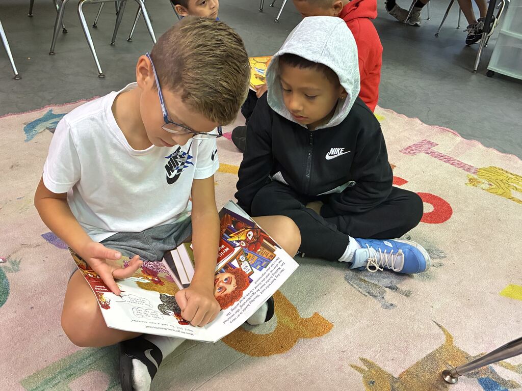 Two boys are sitting on a rug. One reads a book aloud to the other. Behind them, other children sit on chairs.