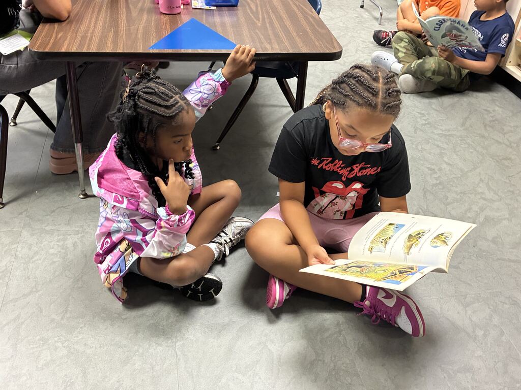 Two young girls sit on the floor, one reading a book and the other listening attentively. Behind them is a wooden table.