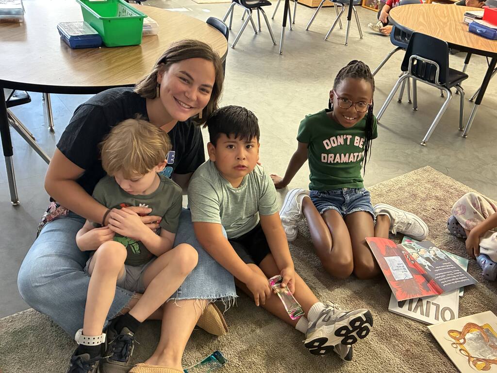 A woman smiles, seated with three children. They are indoors, on a rug. The children are engaged in play. Tables and chairs in the background.