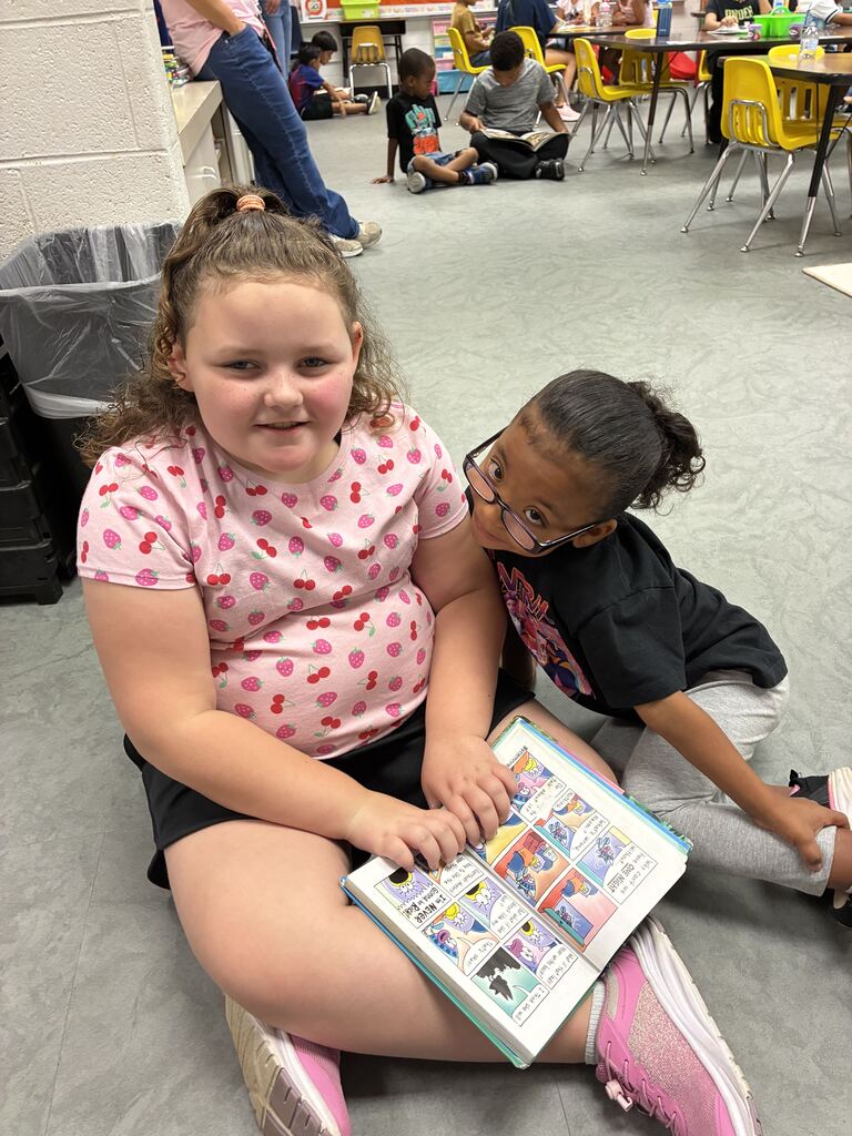 Two children, one with glasses, sit on the floor reading a comic book. Other children are seated or standing nearby in a classroom setting.