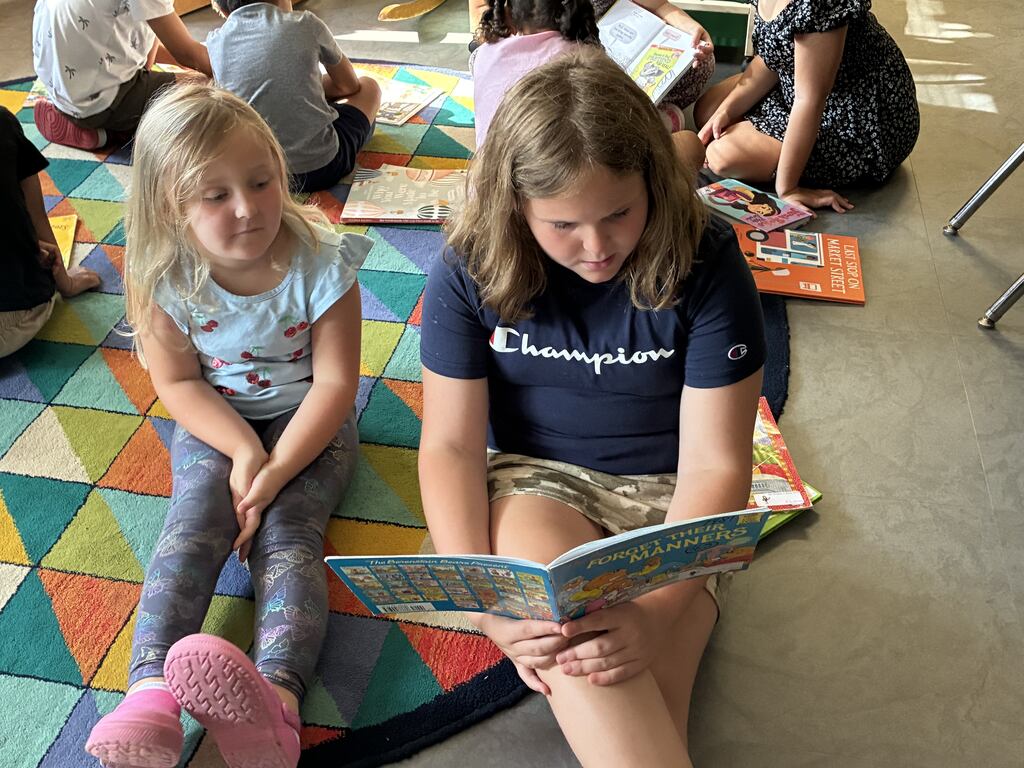 A girl in a Champion shirt reads a book to another girl, both sitting on a colorful rug.
