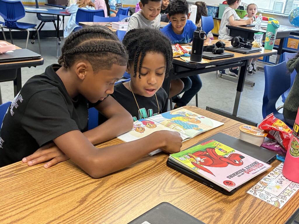 Two children focus on a book at a desk. Other students work at desks behind them. The room has bright lighting and a colorful wall.