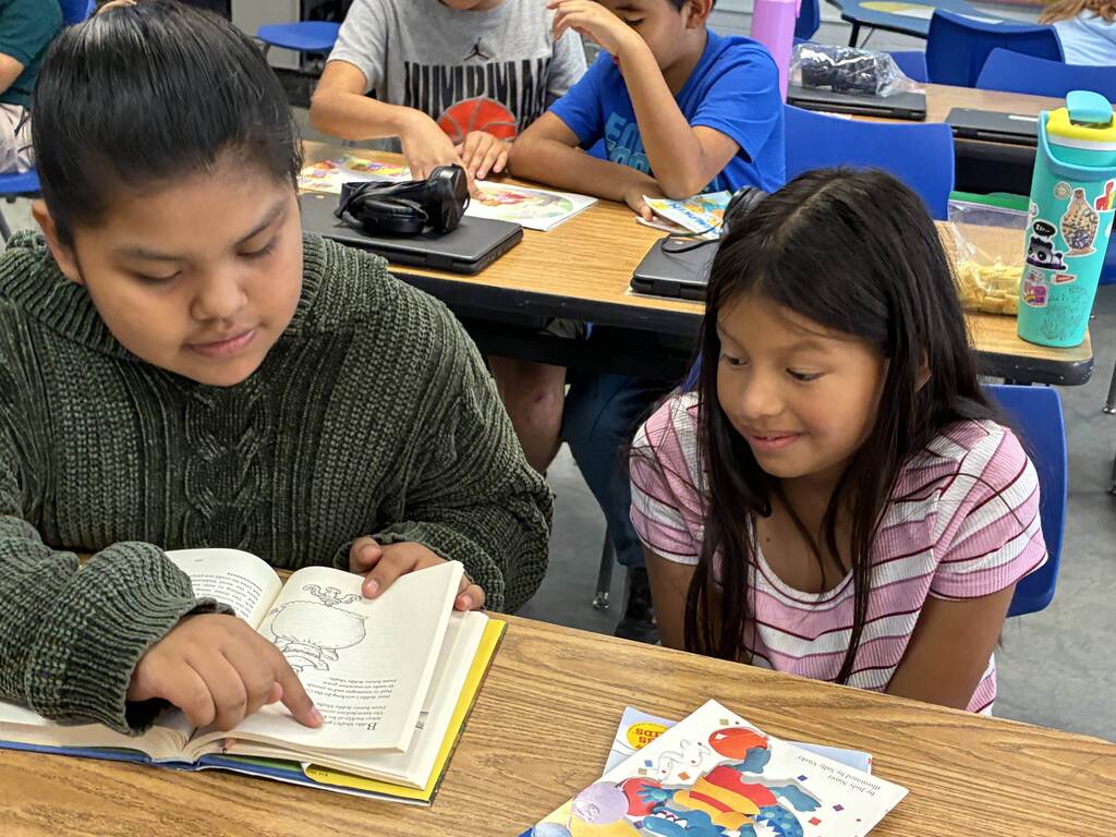 Two girls at desks. One holds a book and points. The other smiles. Background: desks and chairs.