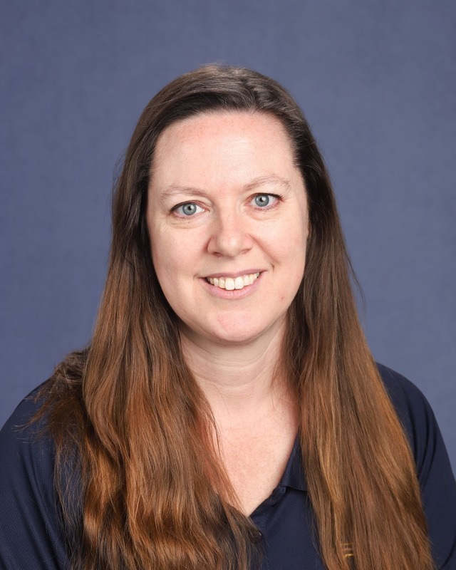 Woman with long wavy brown hair and blue eyes smiling, standing against a solid blue background.