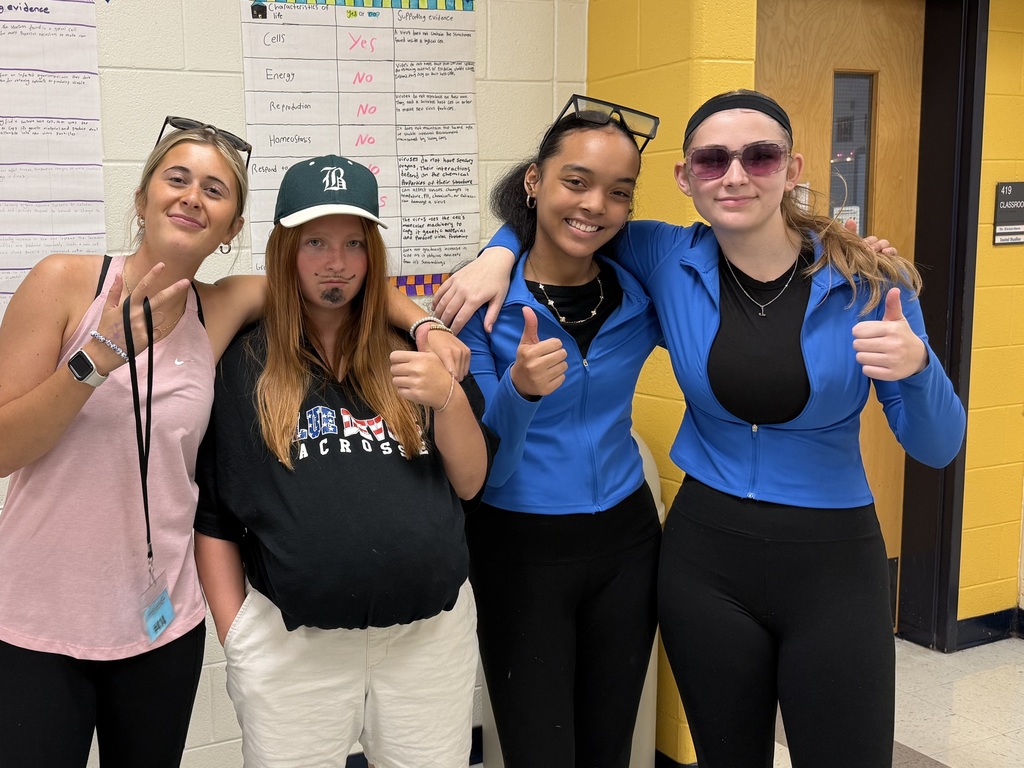 Four women in matching blue tops, black pants, and sunglasses. Two wearing baseball caps. One holds a thumbs-up.