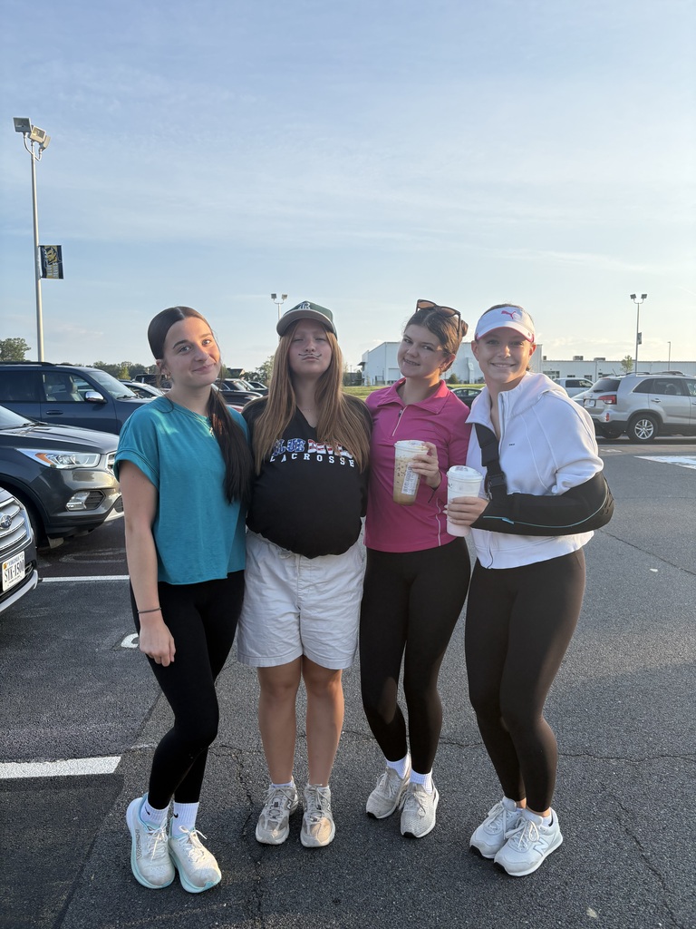 Four women stand in a parking lot, wearing athletic wear. One holds a cup, another has a sling, and the background shows parked cars and street lights.