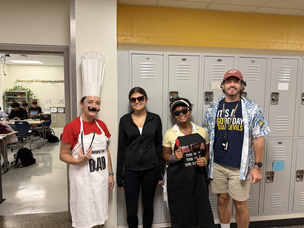 Four people dressed in costumes stand in front of lockers. The background shows a hallway.