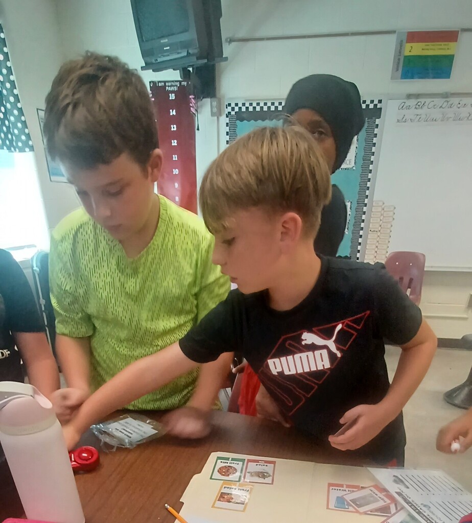 Three boys in a classroom; one in green, one in black, and another in black hijab. They are looking at a table with cards, a pencil, and a water bottle.