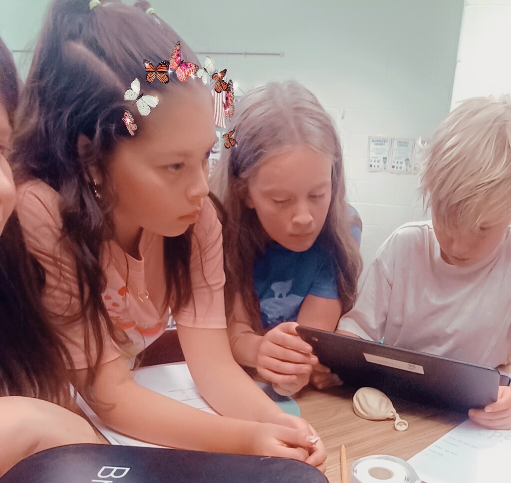 Three children in a classroom. A girl with flowers in her hair, another girl, and a boy with blonde hair.