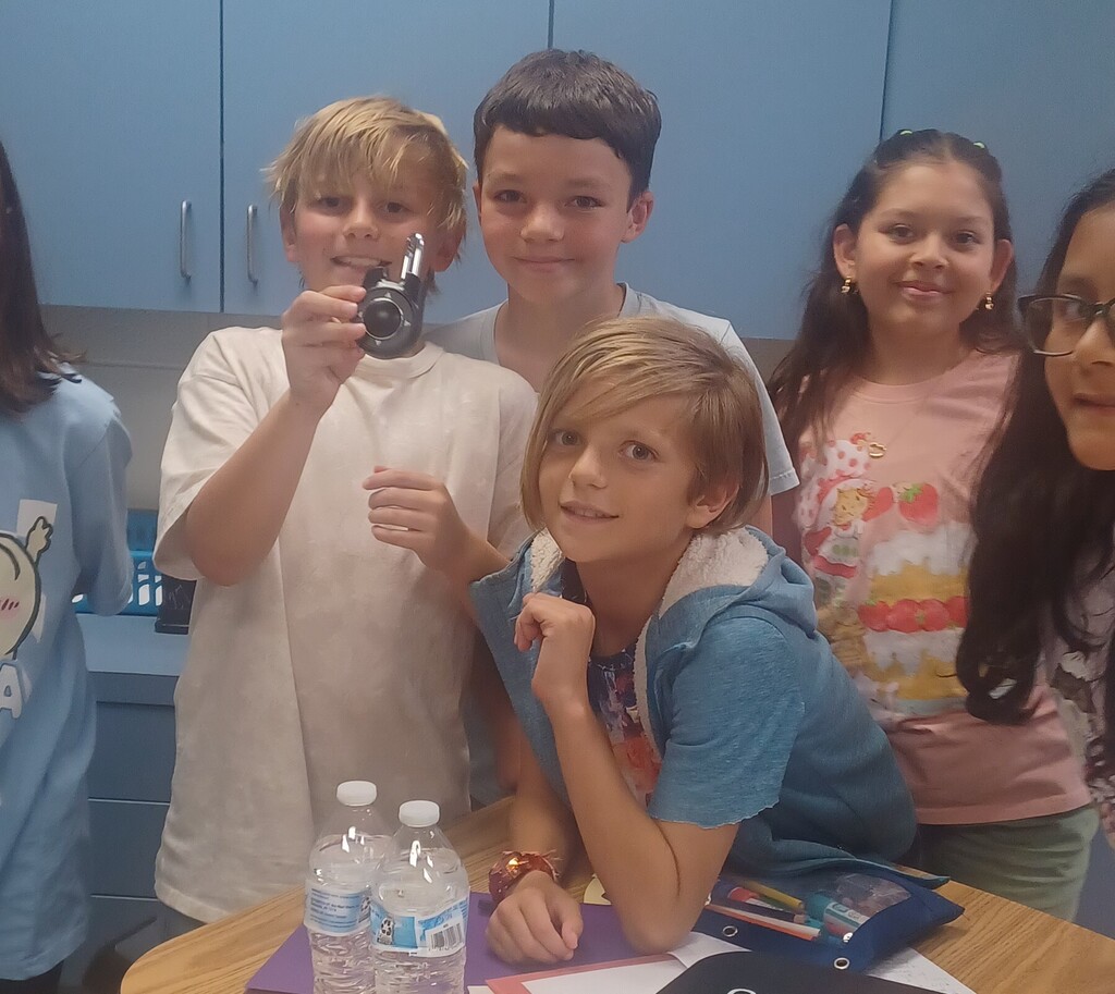 Group of children in a room with blue cabinets, posing for a photo. One boy holds a camera.