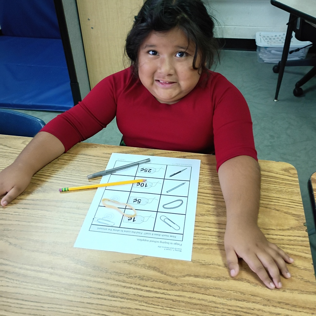 A young child wearing a red shirt sits at a table, holding a paper with shapes and numbers.