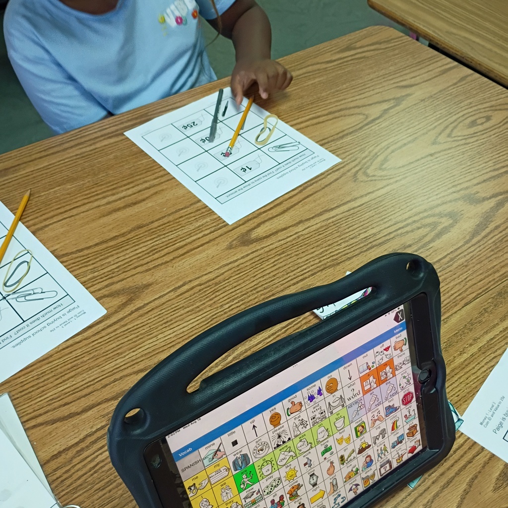 A child in a blue shirt seated at a wooden desk with a tablet and paper in front of them.