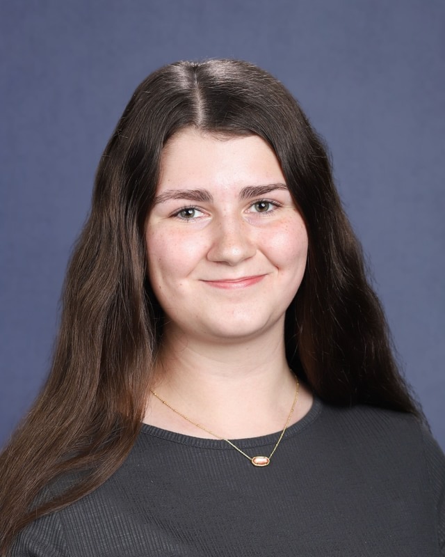 A woman with long hair and a necklace poses against a blue background, wearing a dark shirt.