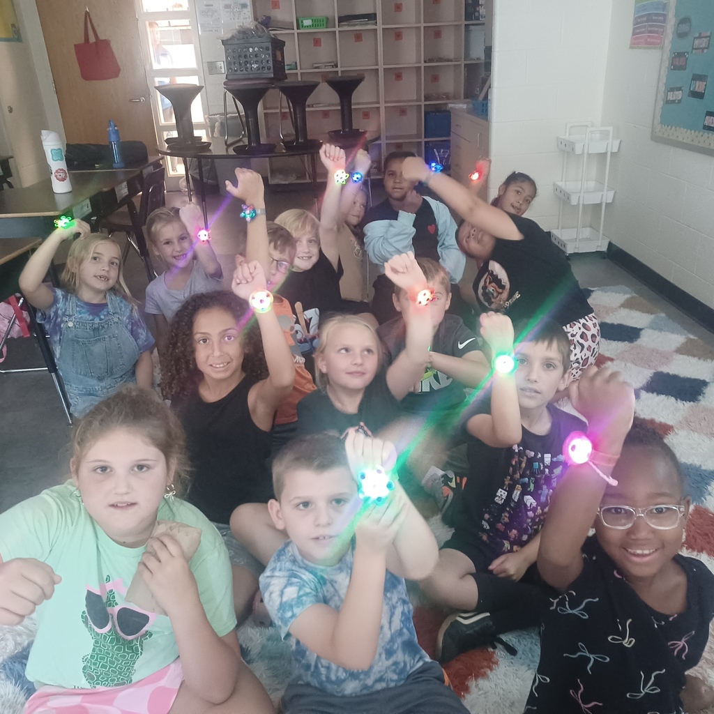Several children sit and crouch in a classroom, wearing glow-in-the-dark bracelets, holding them up.