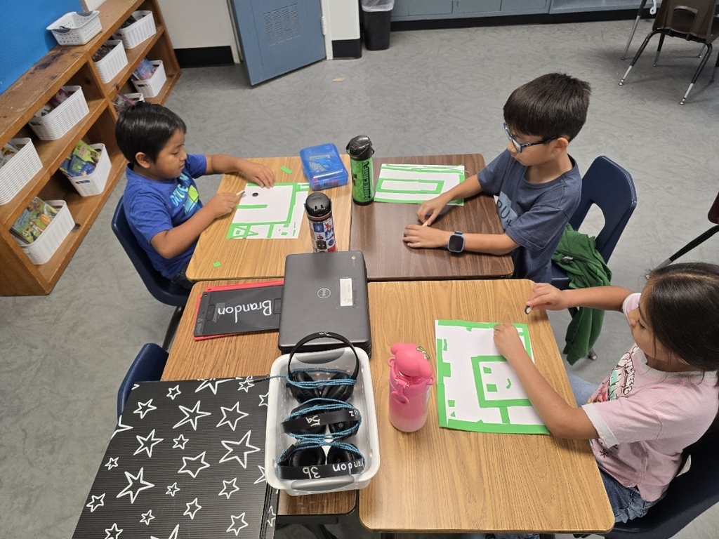 Three children seated at desks with laptops, papers, and a storage box. One child in blue is focused, another in gray is holding a pen, and the third in pink has a pink cup.