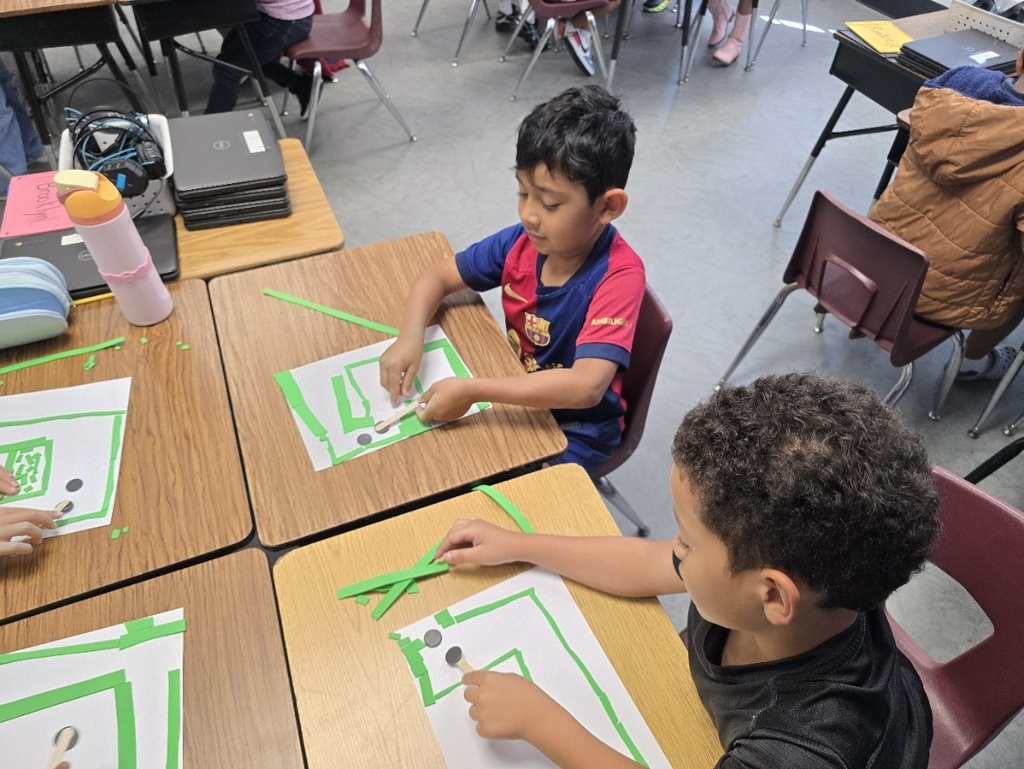 Two young boys in a classroom use scissors to cut green paper into shapes. The desks are covered in white paper with green borders.