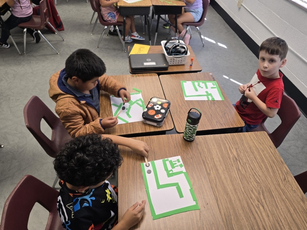 Three children seated at desks, working on a paper with the letter L. One holds a marker.