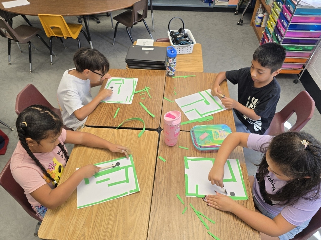 Four children seated at desks, working on green-bordered sheets of paper. One holds a marker.