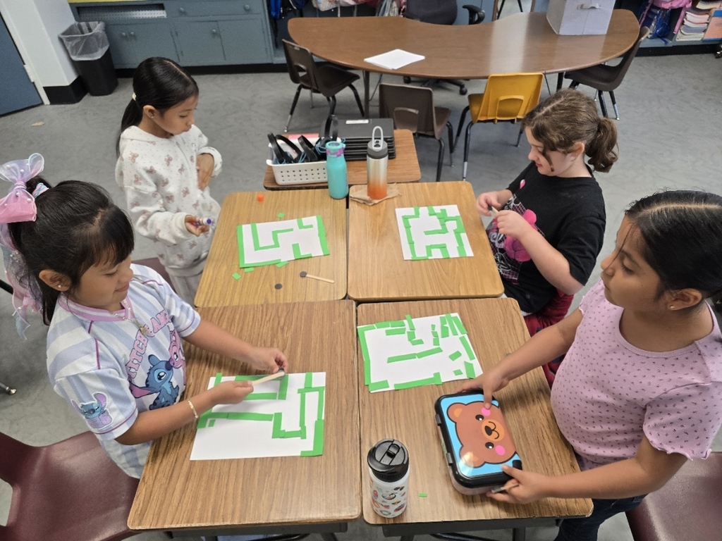 Four young girls seated at desks in a classroom, each engaged with a tablet displaying a puzzle.