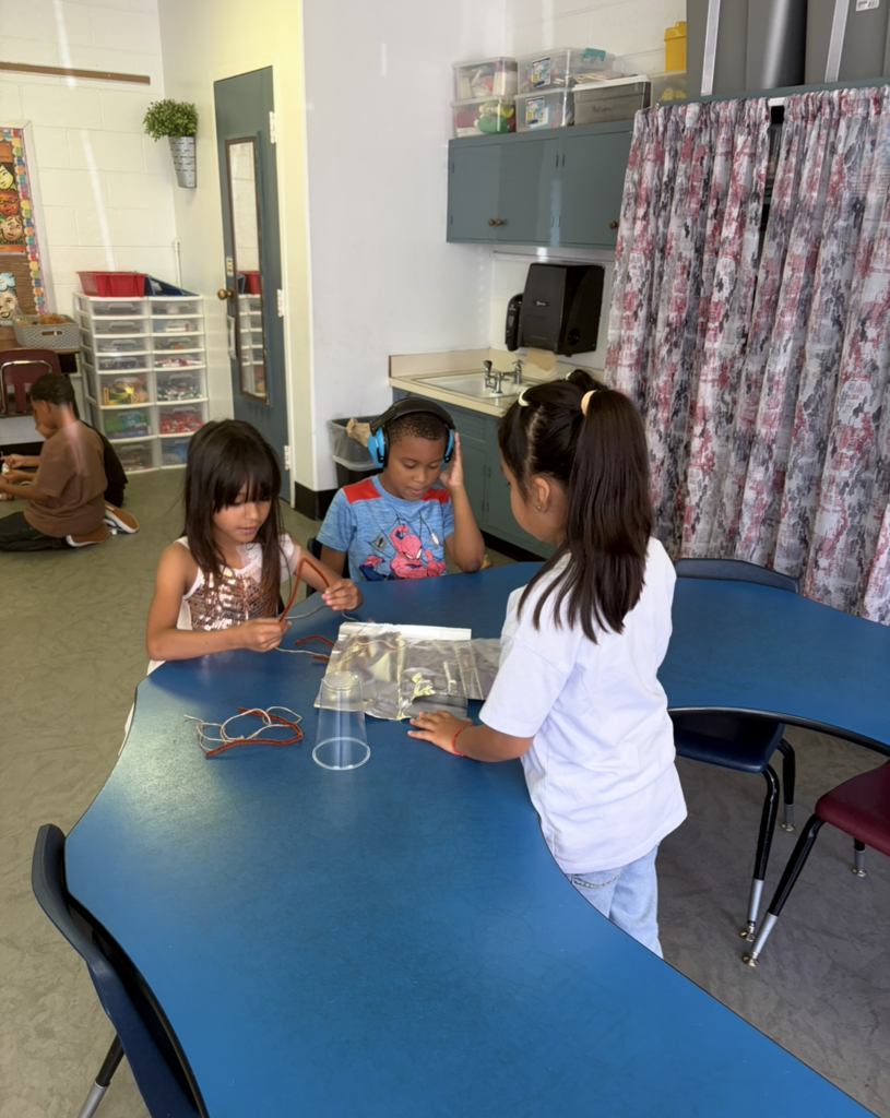 Three young girls in a classroom doing a science project using headphones, wires, and a plastic cup.