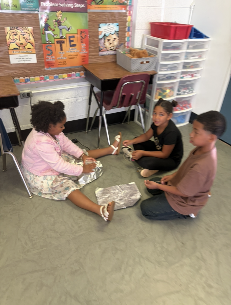 Three children seated on the floor, two girls and a boy. One girl wears sandals and holds paper. Behind them is a classroom with desks, a chair, and a bulletin board.