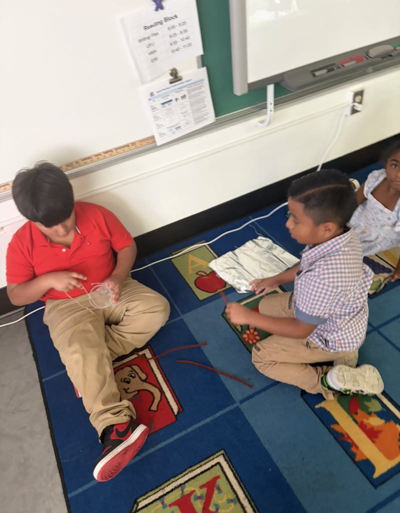 Three children seated on a carpeted floor, one in red, one in blue, working on a project.
