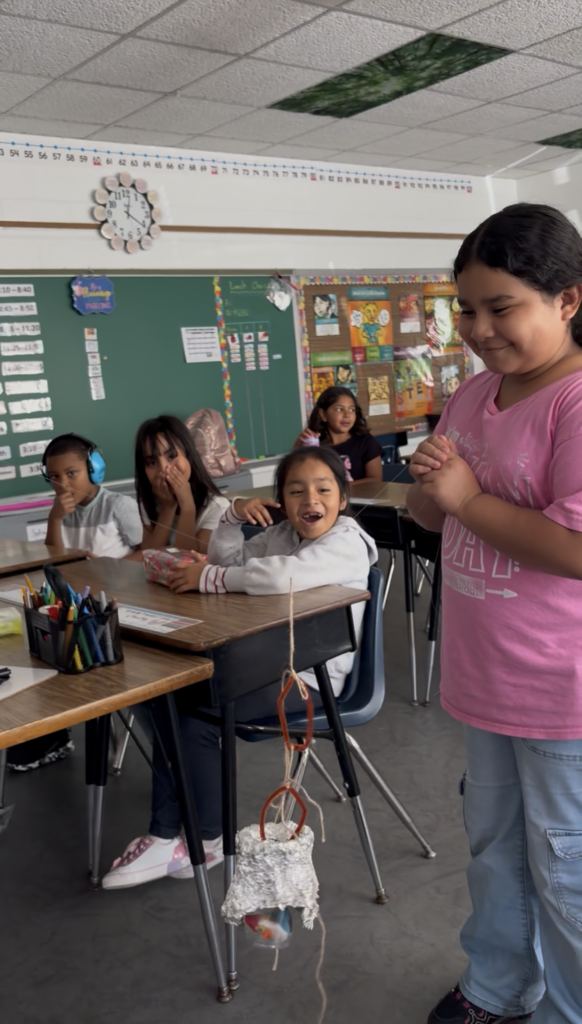Classroom with students seated at desks. One student stands next to a desk with a handmade object hanging from the ceiling.