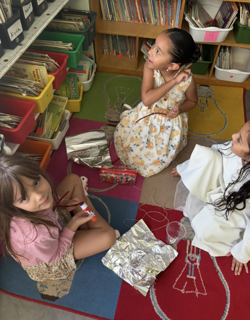 Children in dresses sit on the floor around a rug with a light bulb design. One holds a bottle, and a storage box and books are behind them.